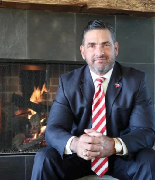 Sam R. Cammack III seated beside a fireplace, wearing a dark suit and red striped tie.